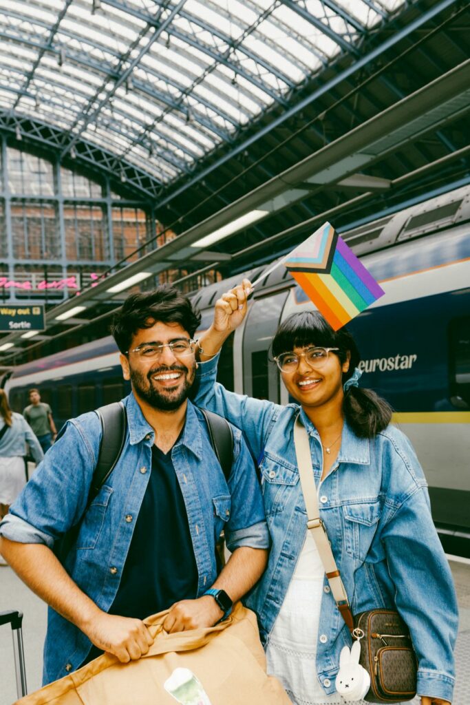 Two smiling people in blue denim jackets wave Pride Progress flags in front of a Eurostar train