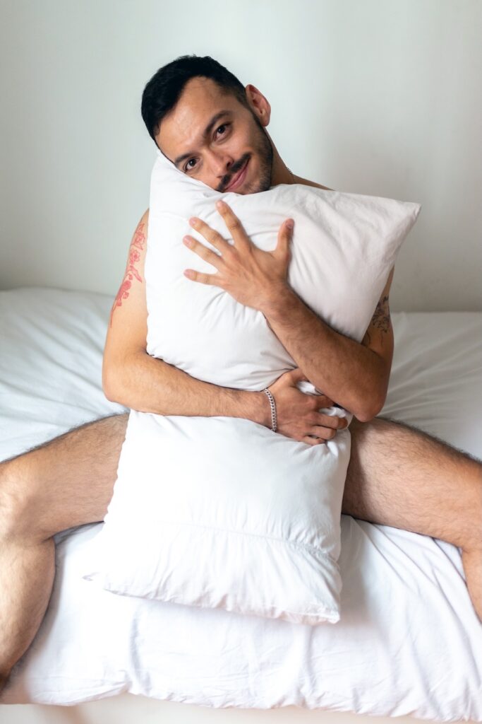 Angel M on a white bed clutching white pillow over body
