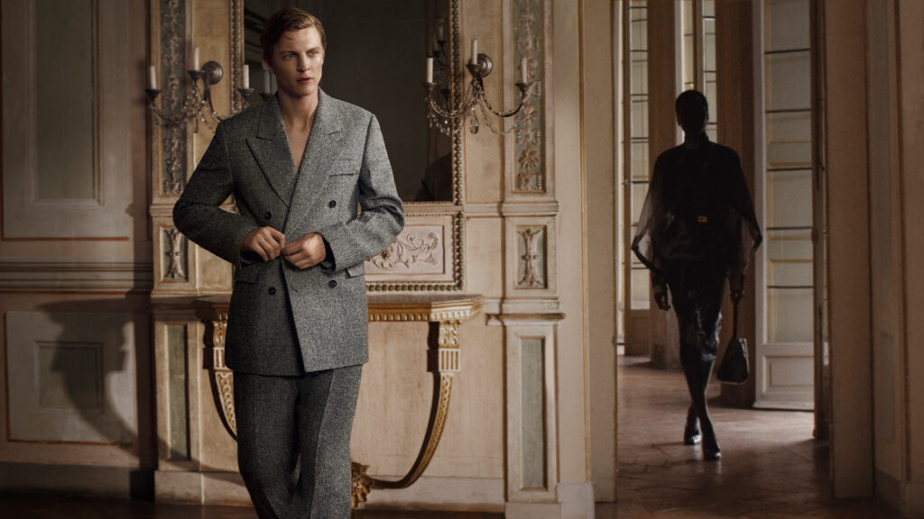 A male model wearing a grey double-breasted suit stands in a dimly lit hallway in Ferragamo's autumn/winter 2025 campaign.