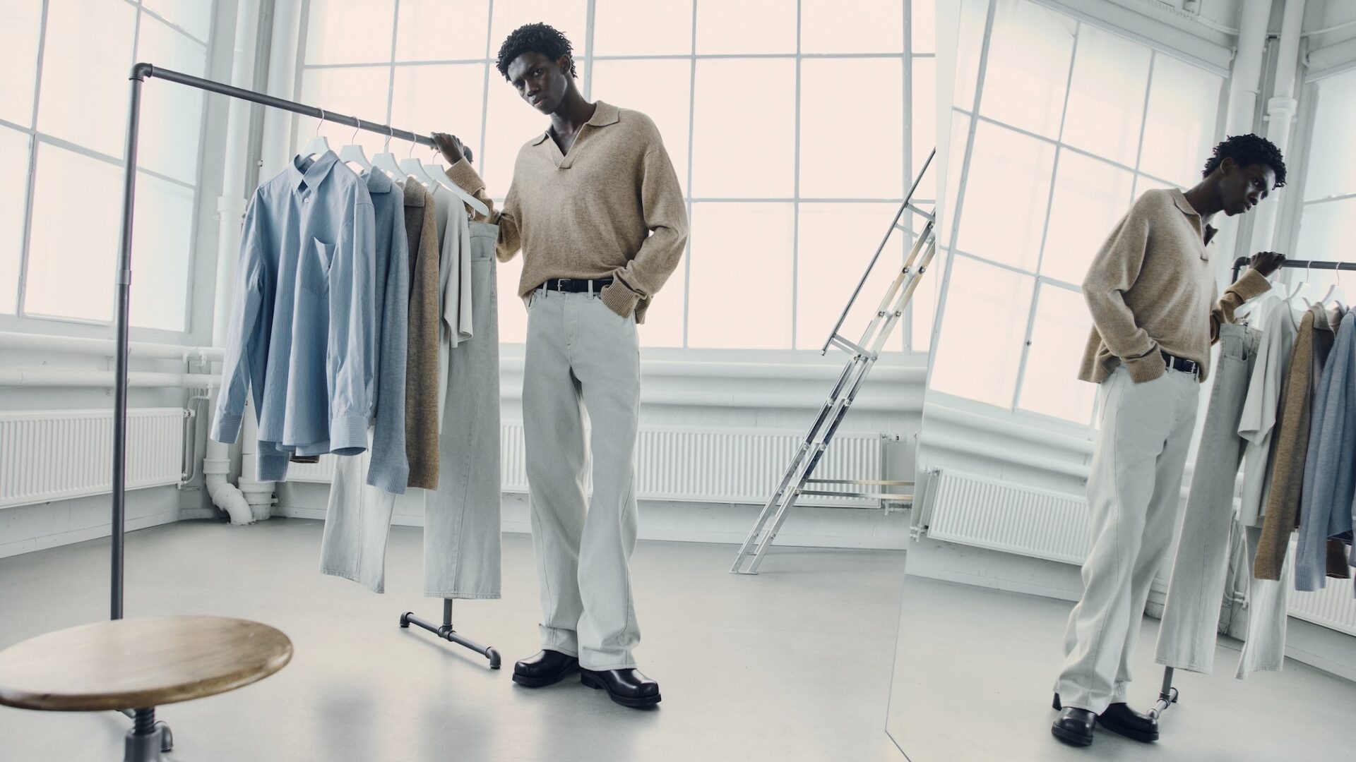 A male model stands in a white, sunlit studio wearing H&M Atelier pre-fall 2025 brown knit and white loose-fit trousers.
