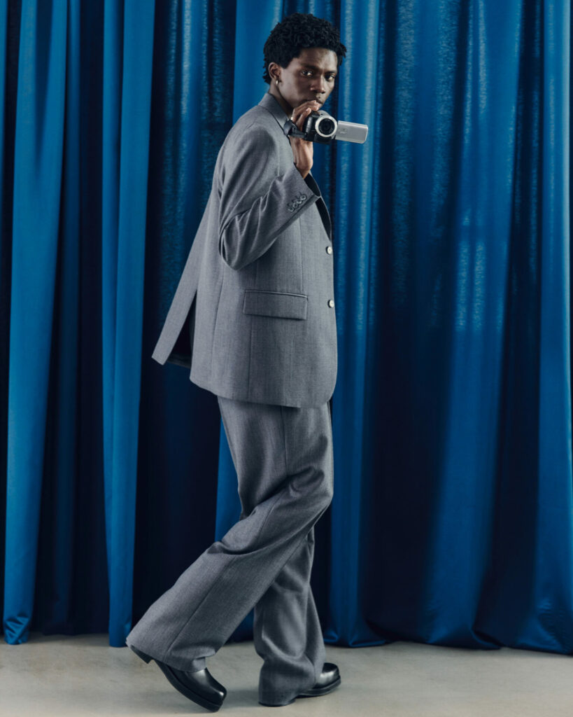 A model stands in front of blue curtains holding a camcorder facing the camera. He wears a slightly oversized suit in grey from the H&M Atelier pre-fall 2025 collection.