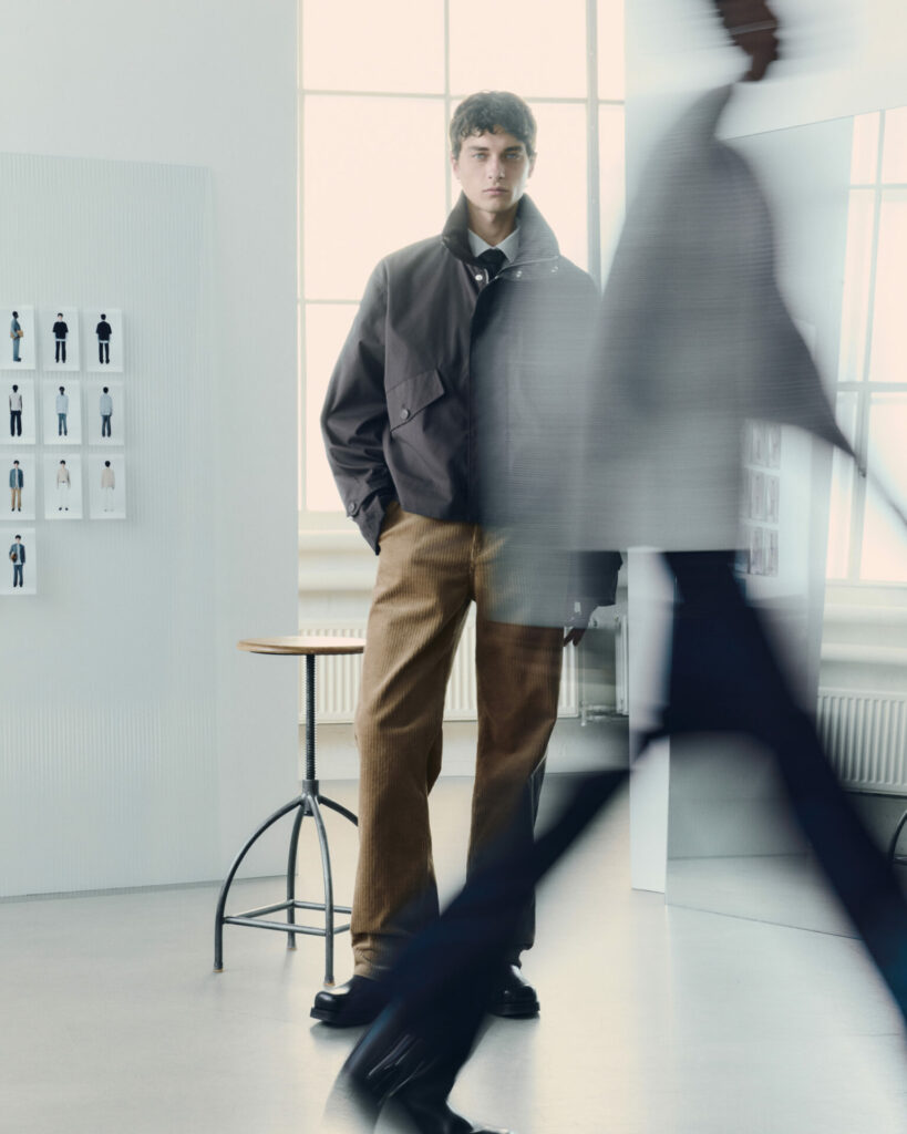 A model stands in a sunlit studio wearing the H&M Atelier pre-fall 2025 collection. He wears a military inspired jacket in grey layered over a shirt and tie.