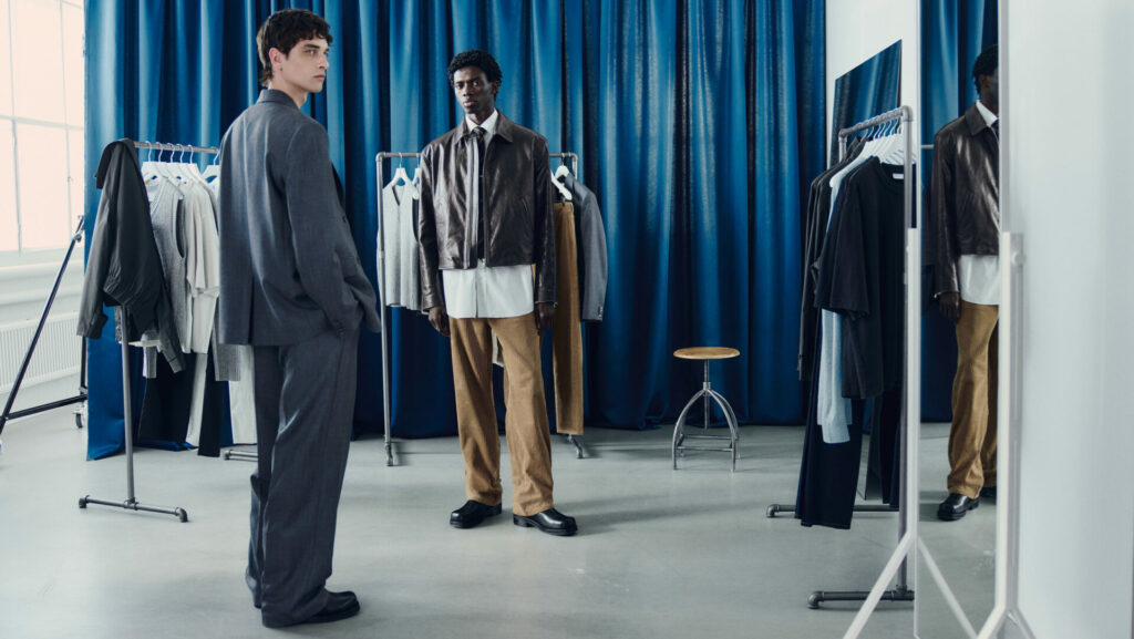 Two models stand in a studio wearing the H&M Atelier pre-fall 2025 collection. One model wears slightly oversized tailoring a cool grey, while the other wears an oversized brown leather jacket layered over an untucked white shirt and straight leg corduroy trousers.