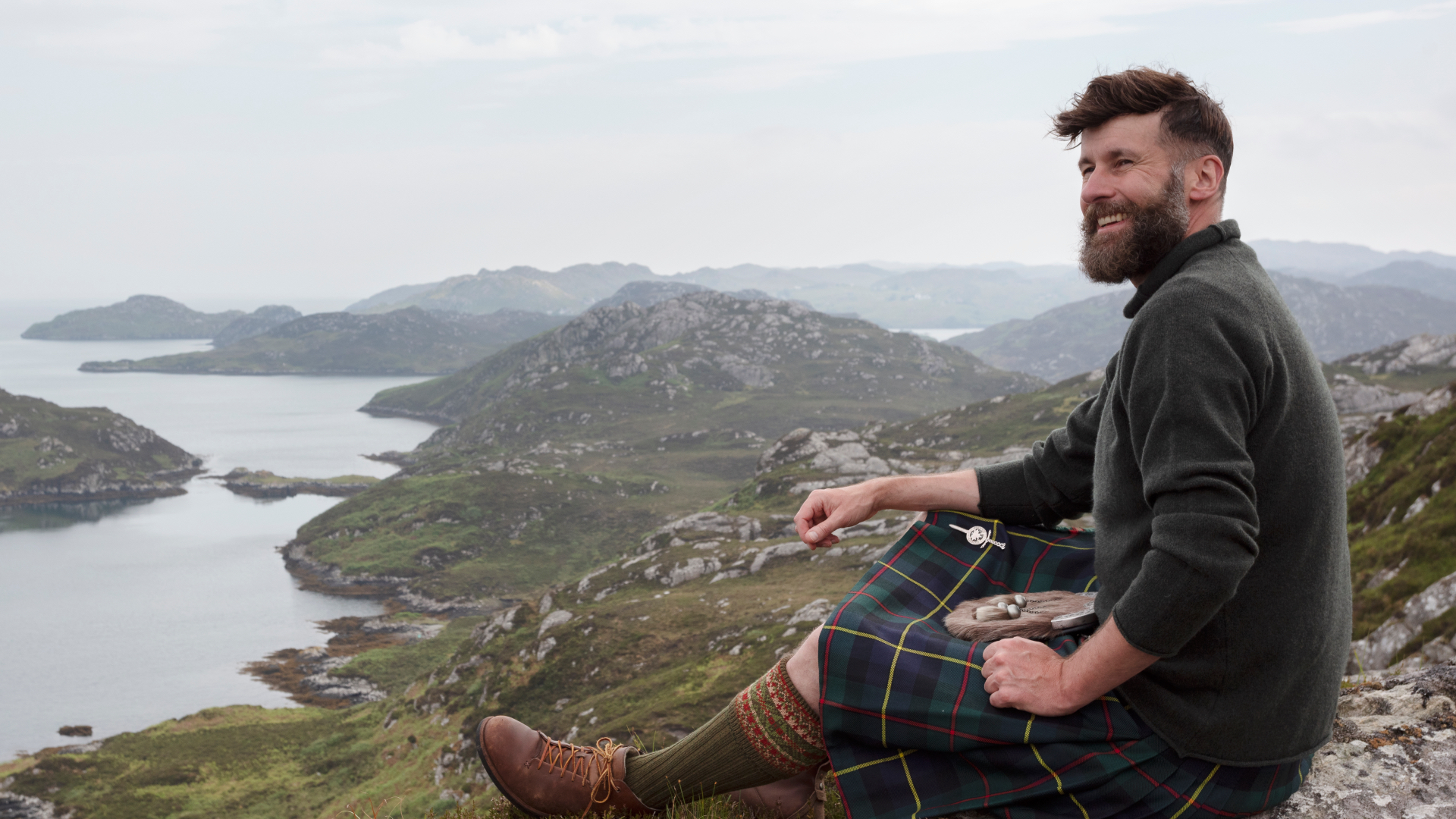 Hebridean Baker in a green jumper and kilt looking out across a scenic mountainous backdrop