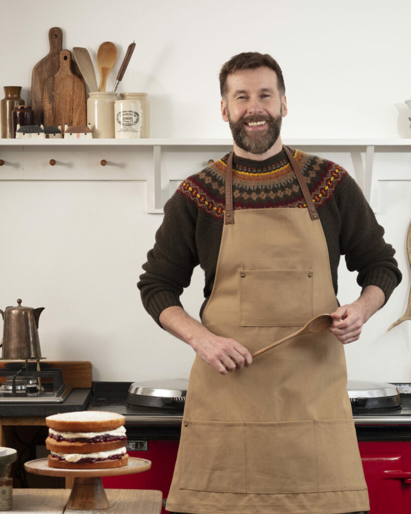 Hebridean Baker in a autumnal coloured patterned jumper covered by a brown apron holding a wooden spoon 