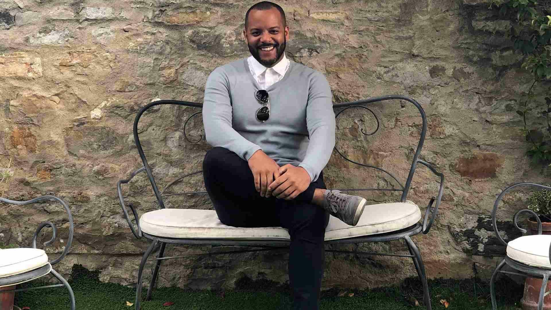 A smiling man sitting on a bench wearing a grey jumper and black jeans