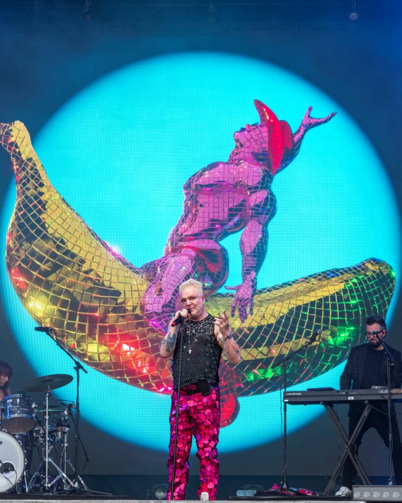 Andy Bell performing on stage at Brighton Pride 2025 with a graphic behind him of a cowboy riding a banana
