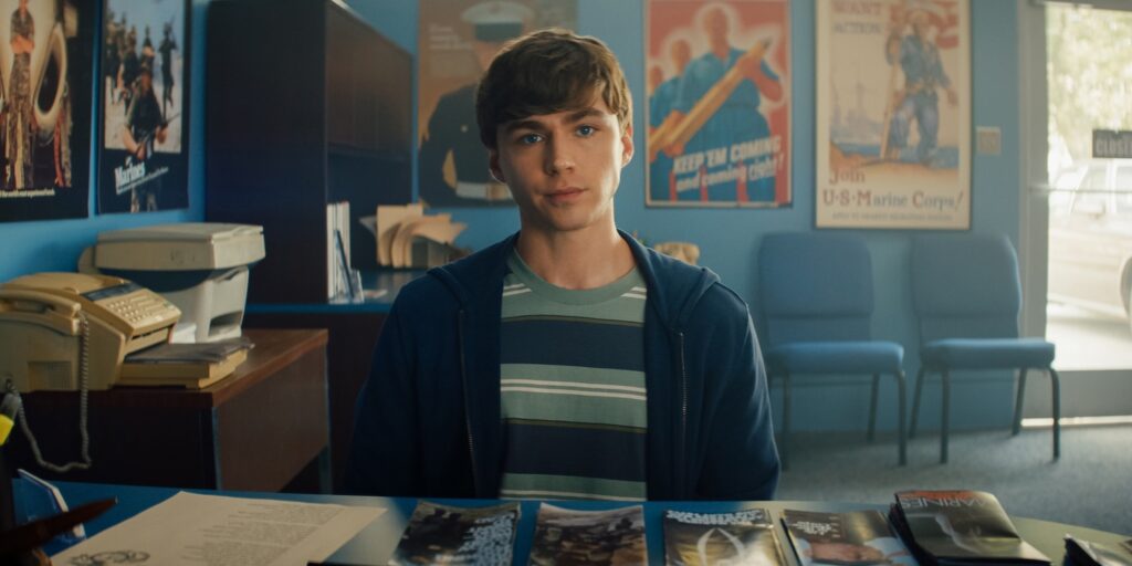 Miles Heizer as Cameron Cope, sat in a blue room and wearing in a striped t-shirt
