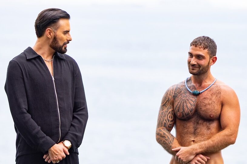 Rylan Clark in a black jacket speaking with a topless tattooed man