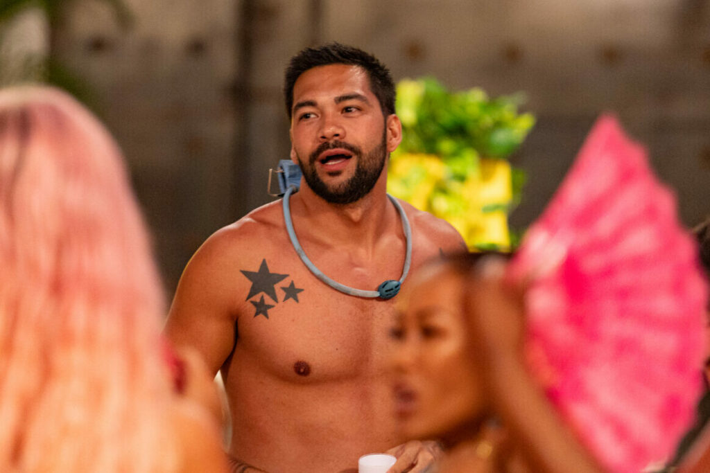 A topless man looking a two women waving around fans