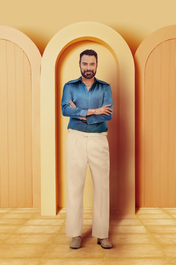 Rylan Clark wearing a turquoise satin shirt and brown trousers with his arms crossed smirking