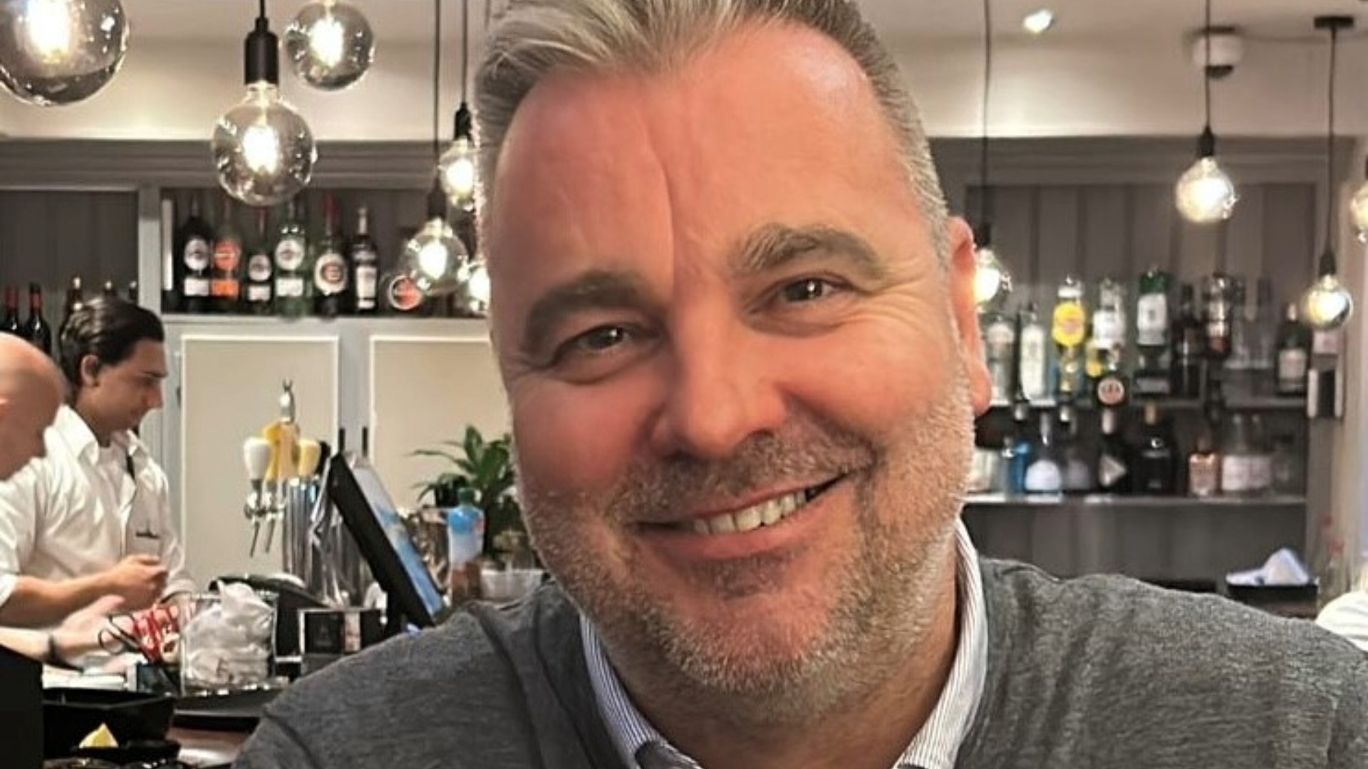 A close shot of a smiling man wearing a grey jumper and white shirt