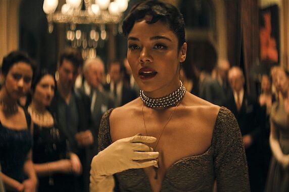 Tessa Thompson wearing a dress and pearl necklace clutching her chest with her white glove dressed hand