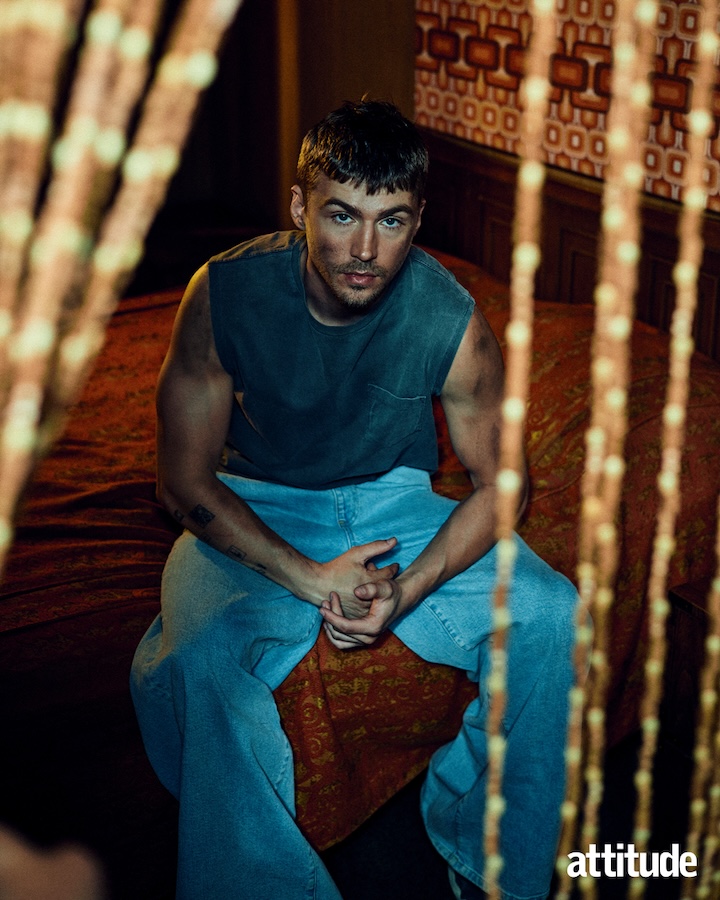 miles heizer in blue tank top and jeans sat on a bed looking at camera