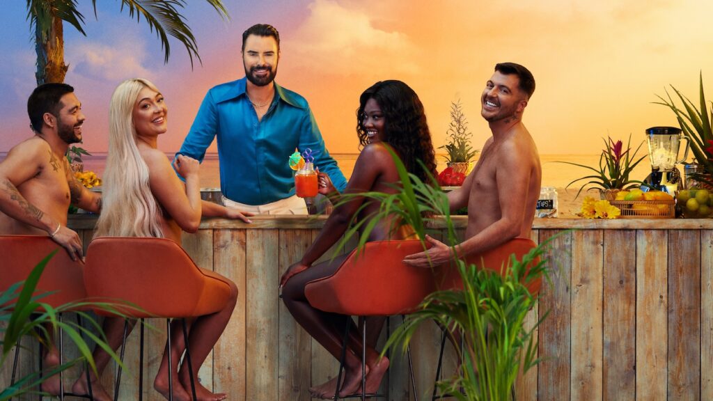 Rylan Clark wearing a turquoise satin shirt leaning on a bar with four naked people sat drinking cocktails