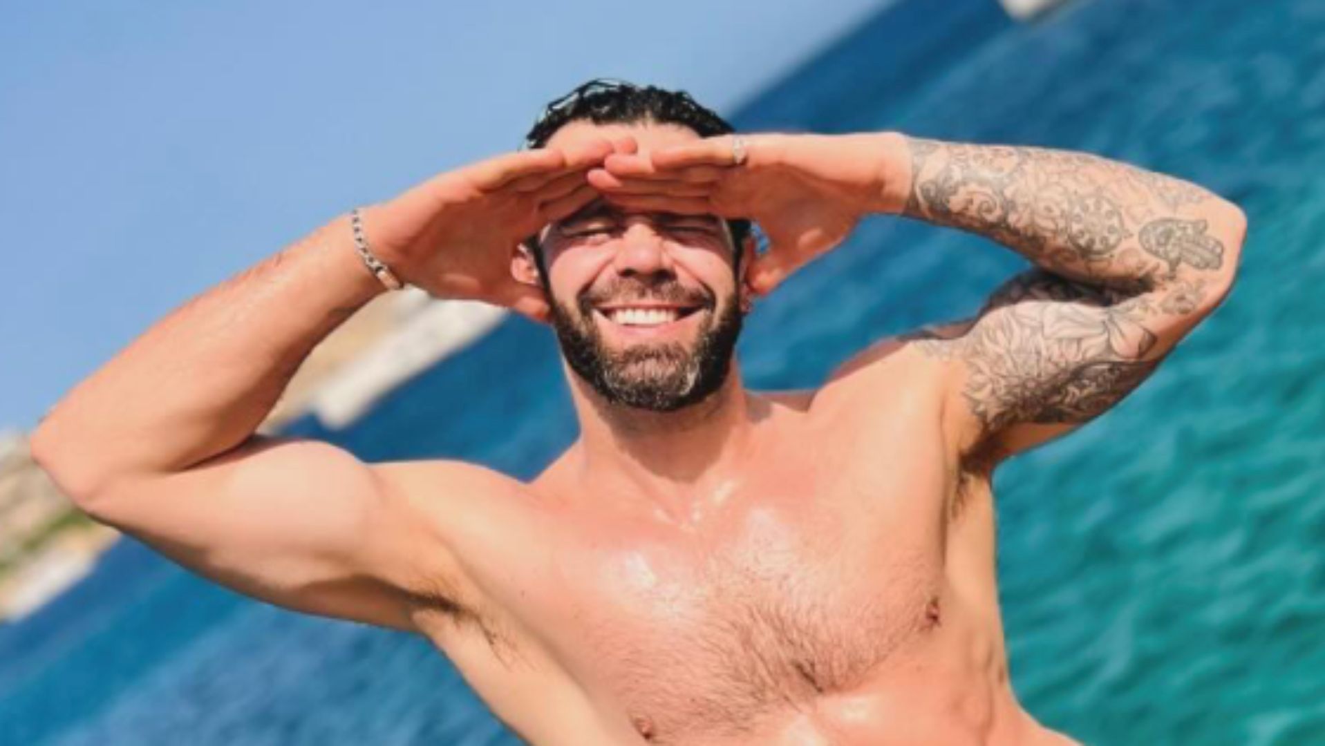Charlie King topless with his arms up flexed by the ocean