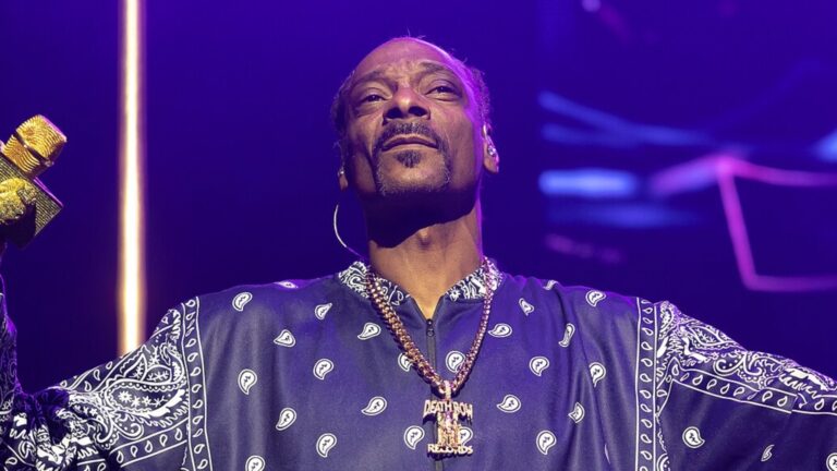 Snoop Dogg in a blue shirt on stage wearing a gold chain holding a gold microphone