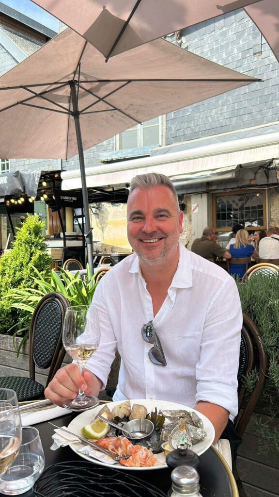 A smiling man in a white shirt holding a glass of wine with a plate of seafood in front of him