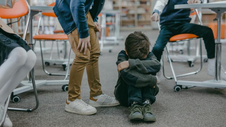 School boy sat on the floor with his head laid in his lap surrounded by people pointing at him