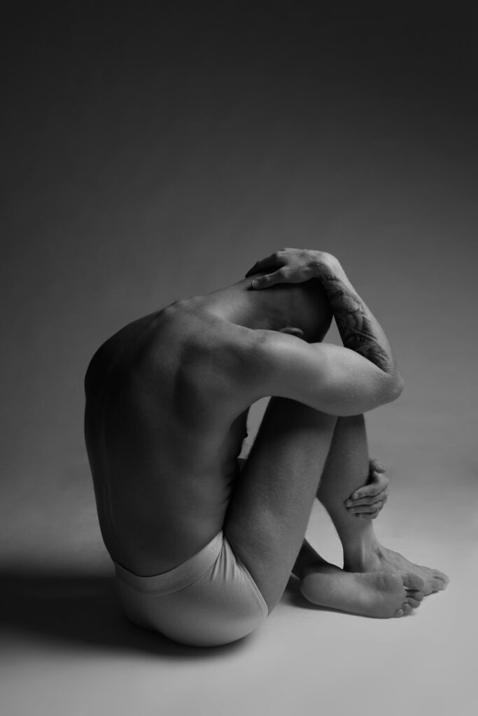 Black and white photo of a man with his head between his legs in just his underwear 