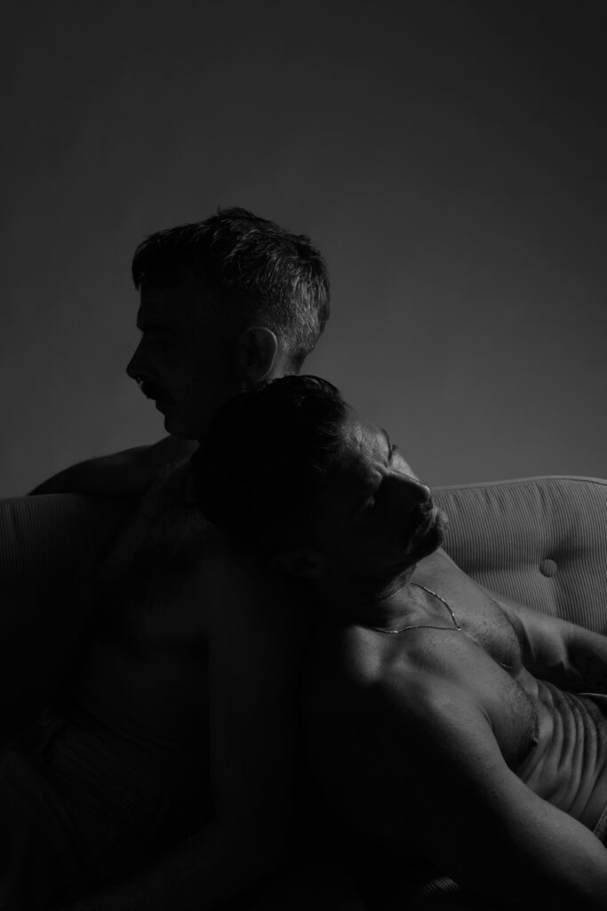 Two topless men leaning up against each other 