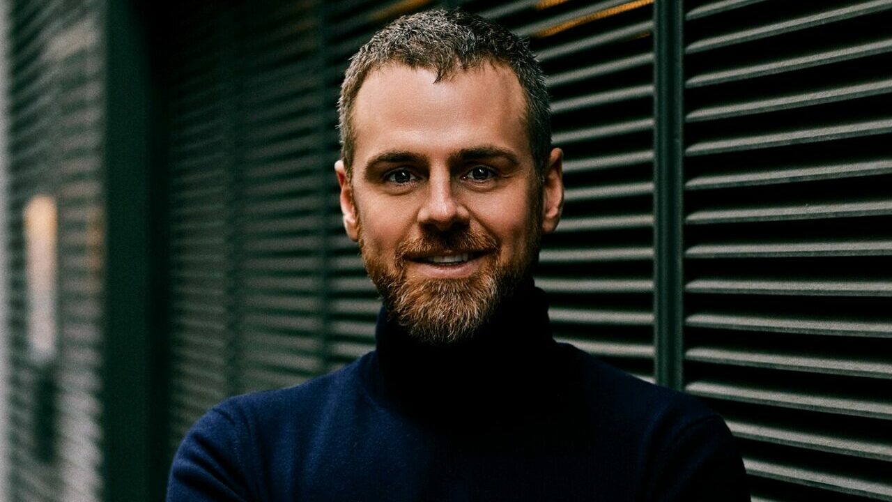 A portrait shot of a smiling man with a beard wearing a dark blue turtleneck jumper