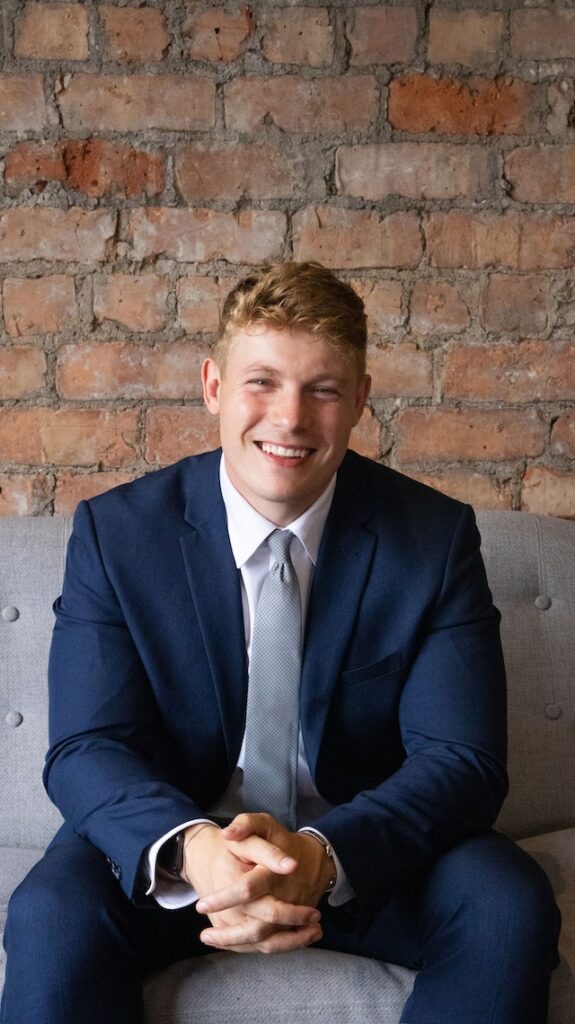 Carl Cashman sitting and smiling in a blue sit and grey tie against with a brick wall behind him