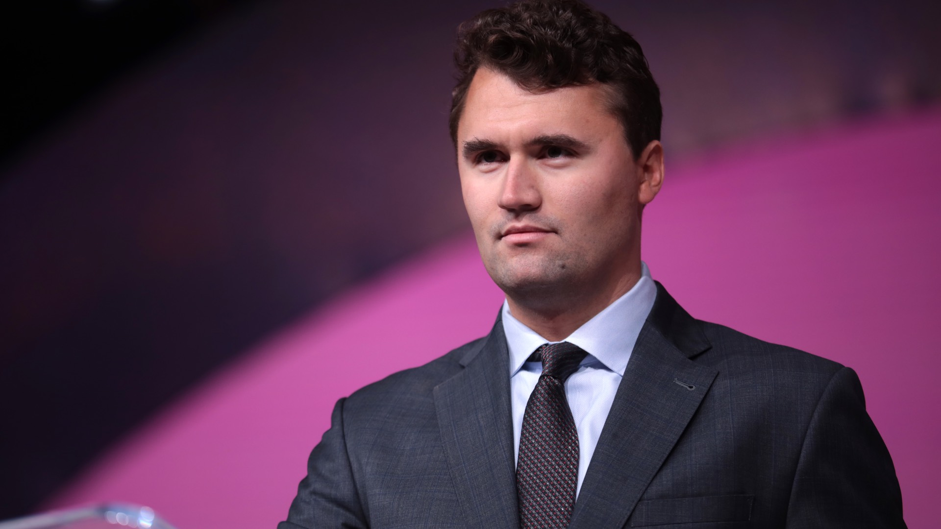 Charlie Kirk in collar and tie, against a black and pink-purple backdrop