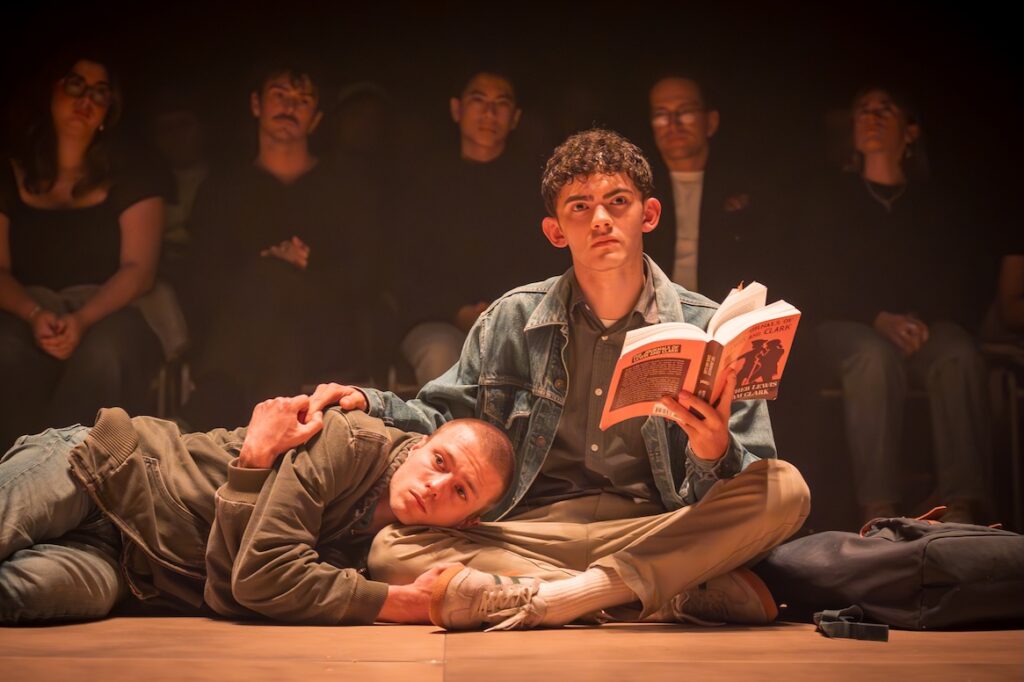 Ruaridh Mollica and Joe Locke in Clarkston, the former lying down, the latter sitting and holding a book, audience members in the background (Image: Marc Brenner)