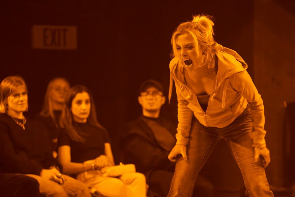 Sophie Melville as Trisha in Clarkston, orange light, she is screaming as audience members look on behind her (Image: Marc Brenner)