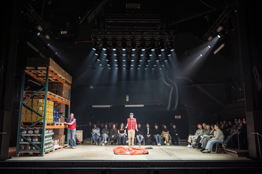 a shot of the stage with the actors visible and audience members sat behind them, a shelving unit on the left