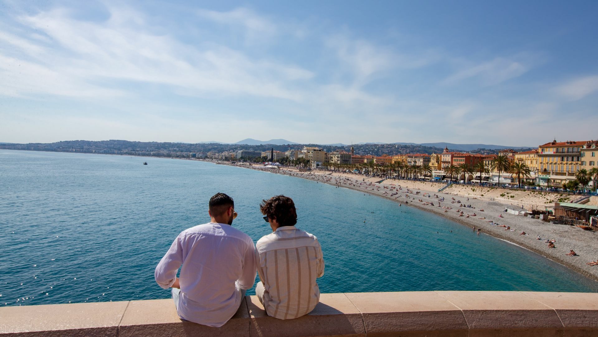 Two men sitting on a wall overlooking the water and buildings of Nice, France