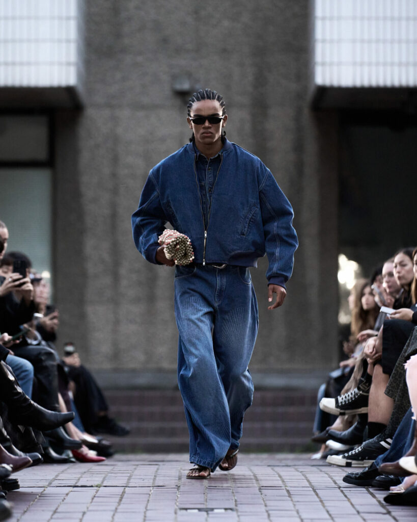 A model walks a fashion show at the Barbican Centre wearing a denim bomber jacket and blue jeans.