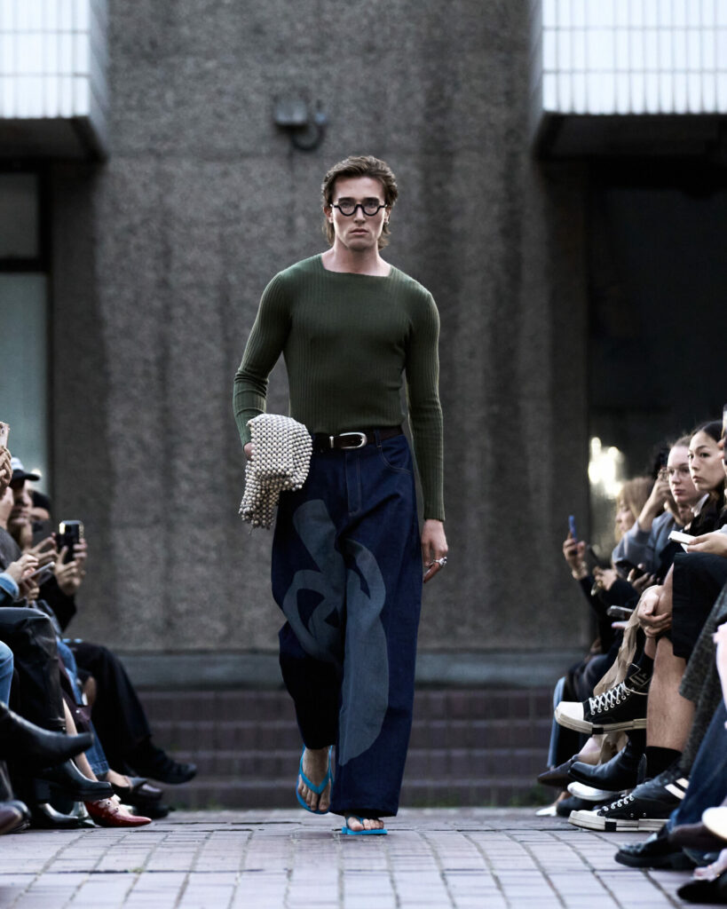 A model walks a fashion show at the Barbican Centre wearing a skintight green knit and wide-leg jeans. 