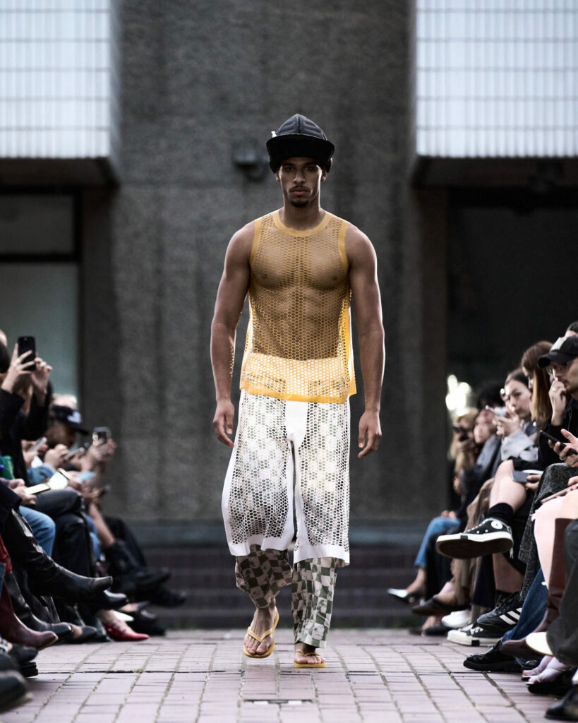 A model walks a fashion show at the Barbican Centre wearing a yellow latex mesh vest.