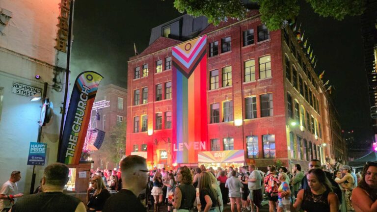 Night view of outside the Leven hotel in Manchester with a giant Pride flag hanging down the front and people walking in front of it