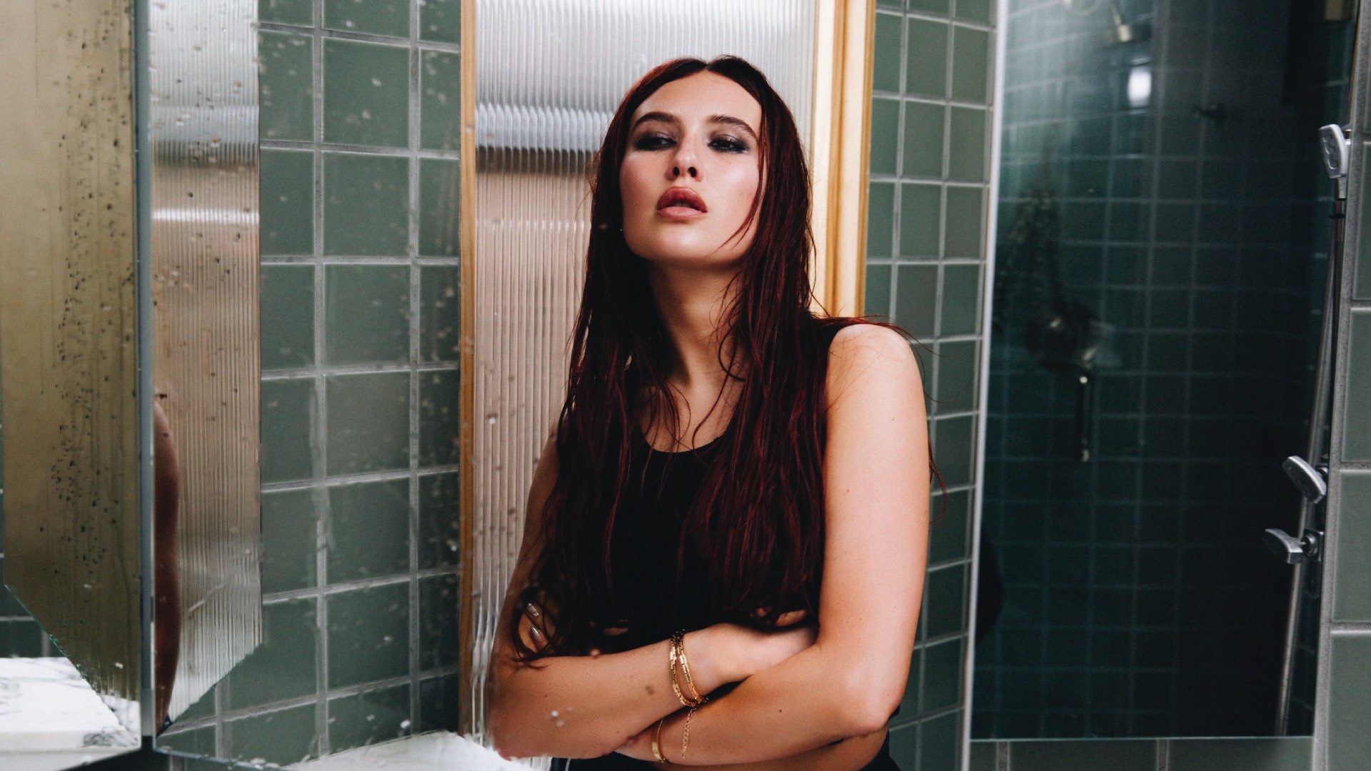 Mimi Webbe with dark hair and black top standing in a green tiled room