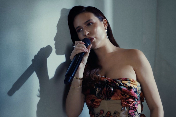 Mimi Webb leaving against a grey wall holding a mic, dark hair, flowery top