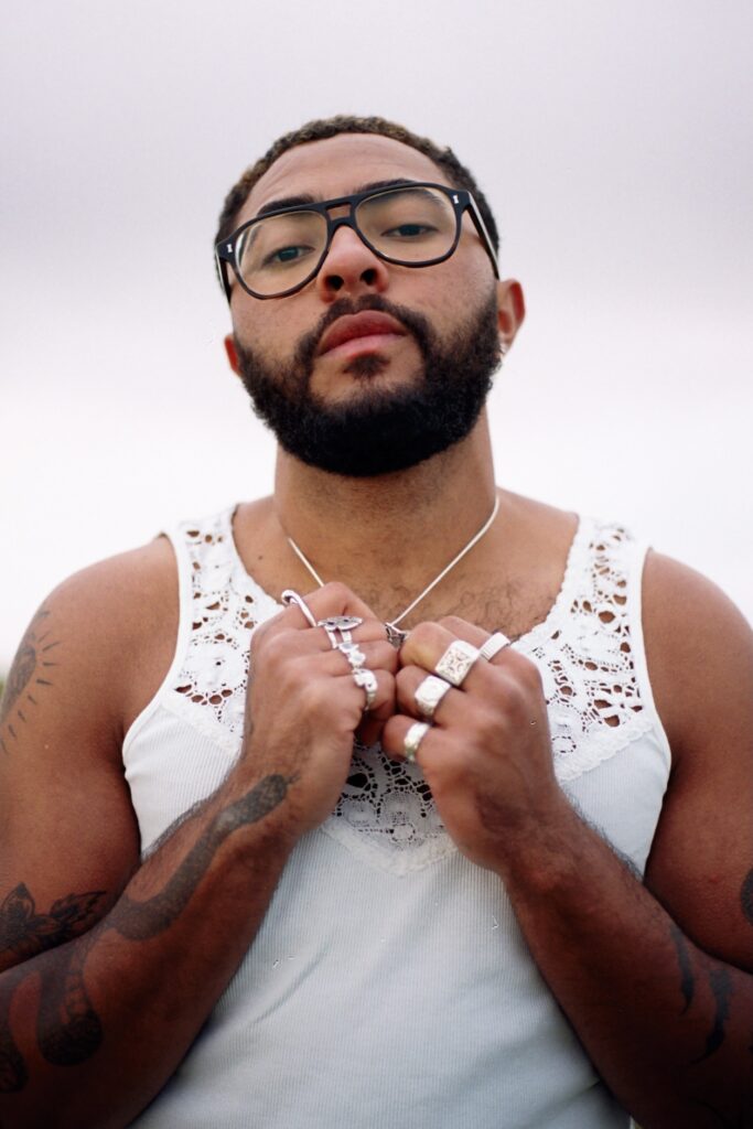 Miri in a white vest with lace at the top, several rings, glasses and beard