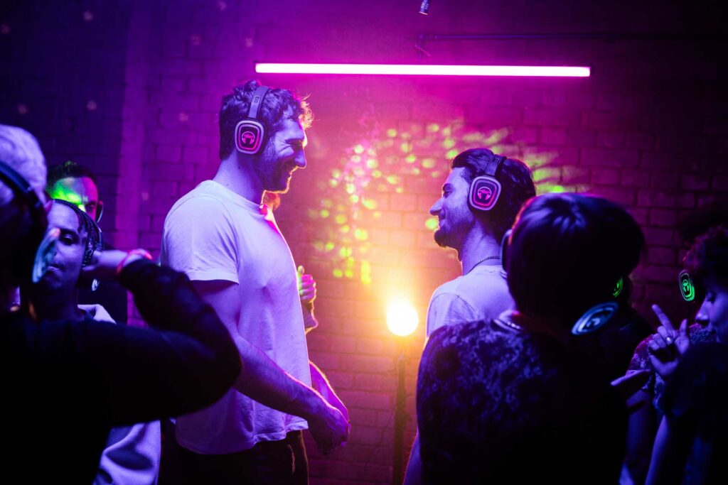 A still from the film A Night Like This showing the two male characters standing in a nightclub wearing colourful headphones