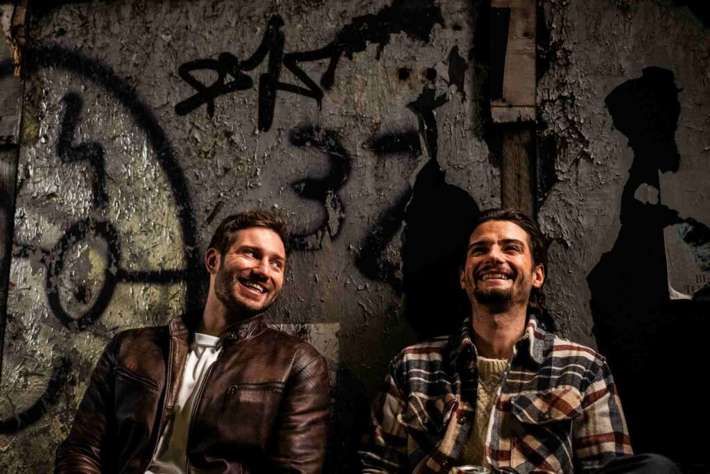 A still from the film A Night Like This showing the two male characters sitting in front a graffiti covered wall