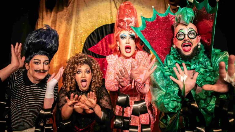 Four drag performers in flamboyant oufits posing on a theatre stage