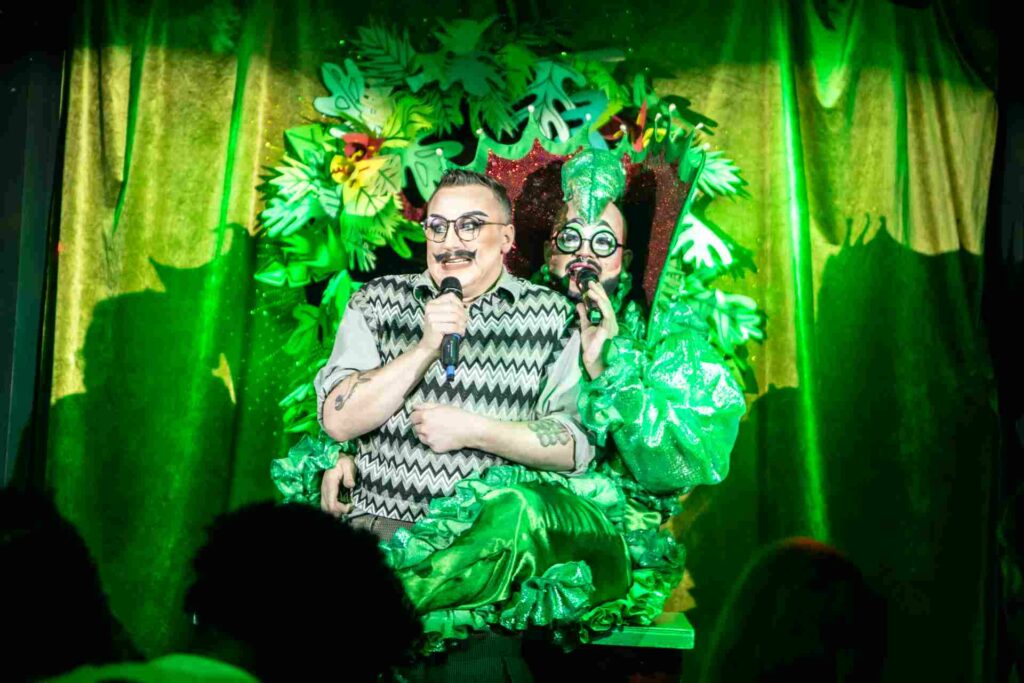 Fatt Butcher and a fellow performer in green theatrical costumes singing on stage