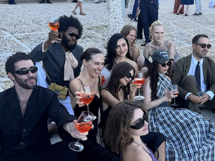 A group of celebrities surrounding Charli XCX in her wedding dress rasiing glasses of Aperol spritz 