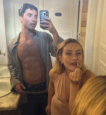 Benito Skinner with Amelia Dimoldenberg applying lip gloss in a bathroom mirror 