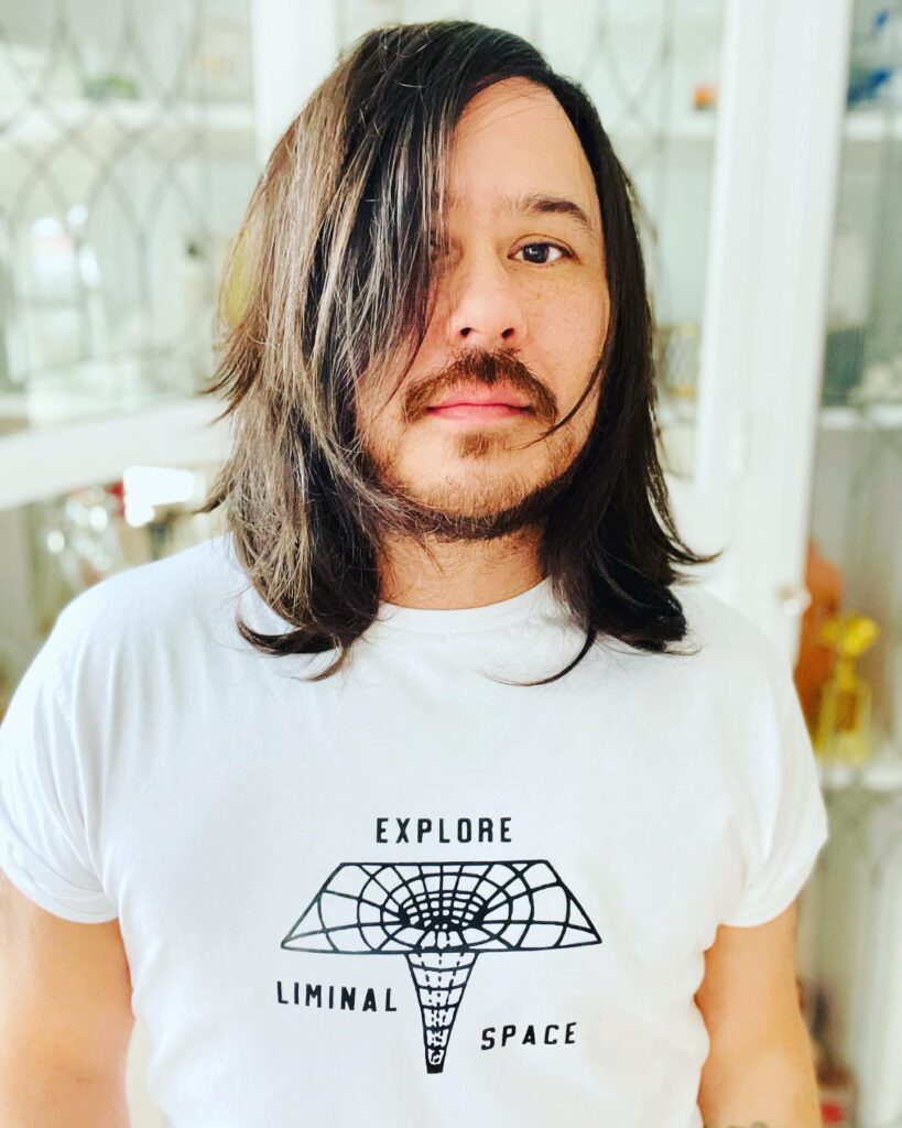 Theodore with long brown hair and beard, white t-shirt with words 'explore liminal space' on it