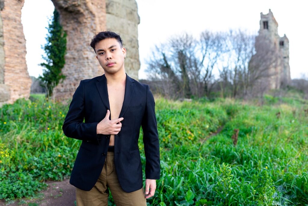 andrewson M in a black jacket and brown trousers standing by some grass and some Roman ruins
