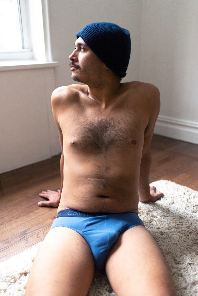 Dave P sitting down in blue underwear on a rug and wooden flooring wearing a blue hat, topless