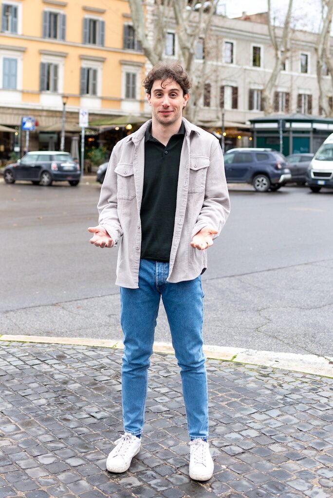 Leonardo F standing in the street with blue jeans on, black t-shirt, white jacket, holding his hands up