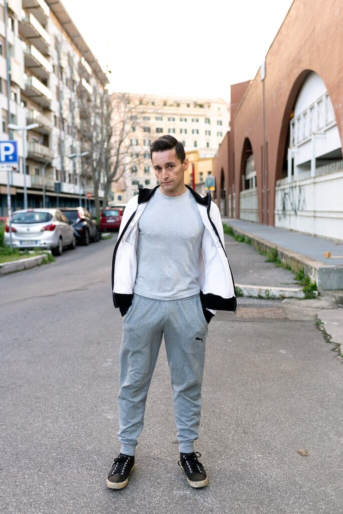 Lucas G in grey sweatpants and black trainers and grey t-shirt and white jacket standing on a street